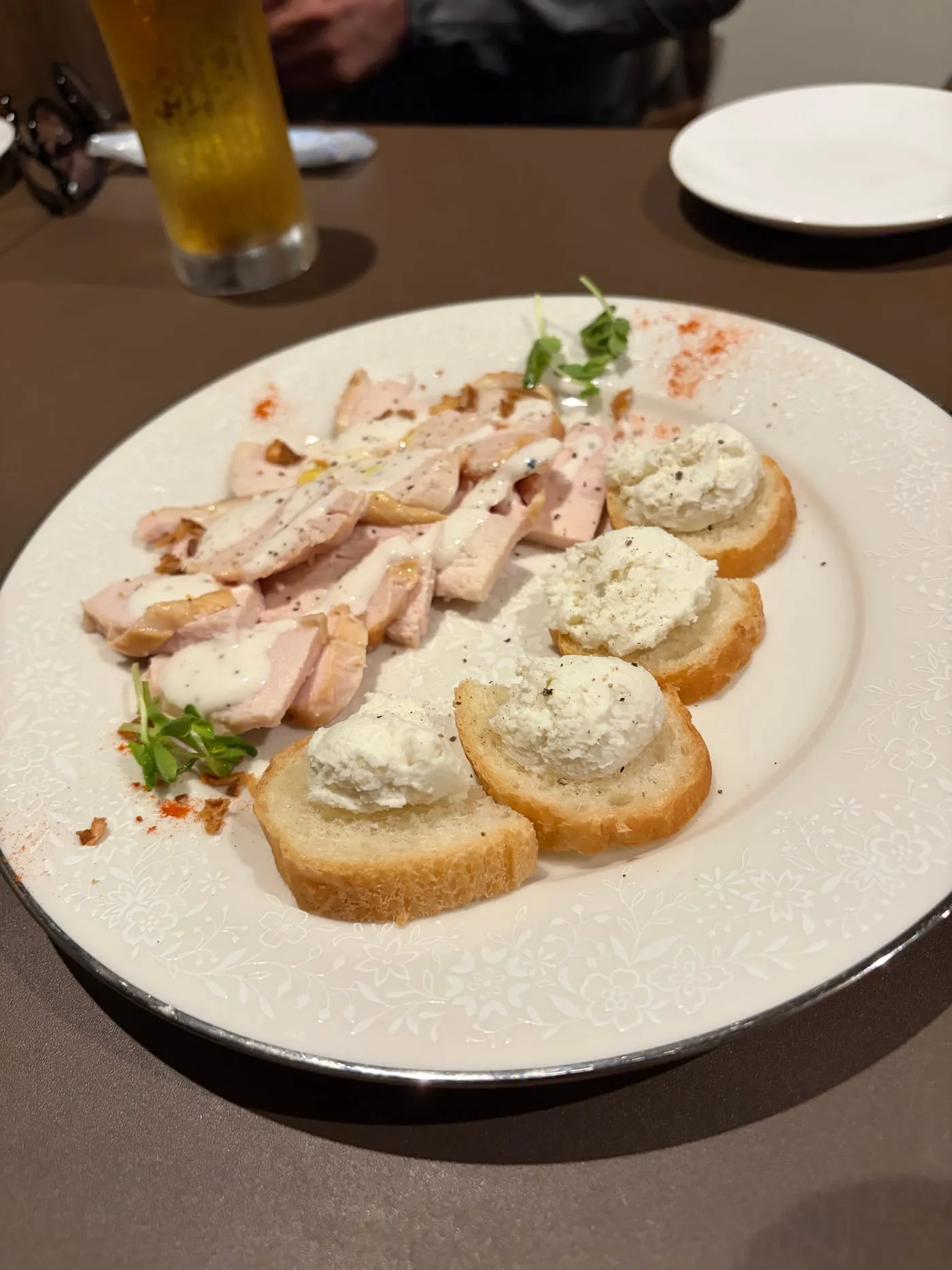 白い皿に盛られた前菜。スモークした鶏肉の薄切りに白いソースがかかり、奥にはバゲットの薄切りが並んで、それぞれに白いクリームチーズが乗せられ黒こしょうがふってある。手前に小さな緑の葉が添えられている