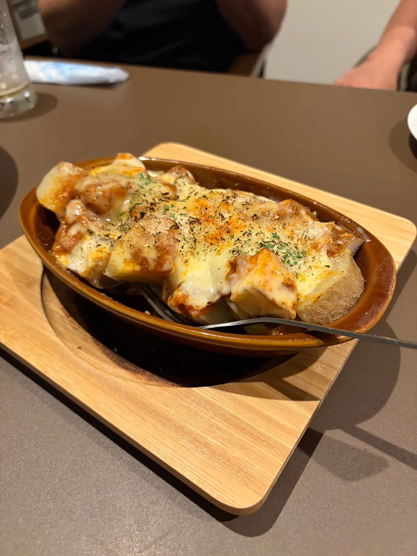 木の板に乗せられた茶色いグラタン皿。角切りの具材にとろけたチーズがかかって焼かれ、パセリと粉チーズがふられている。スプーンが添えられている