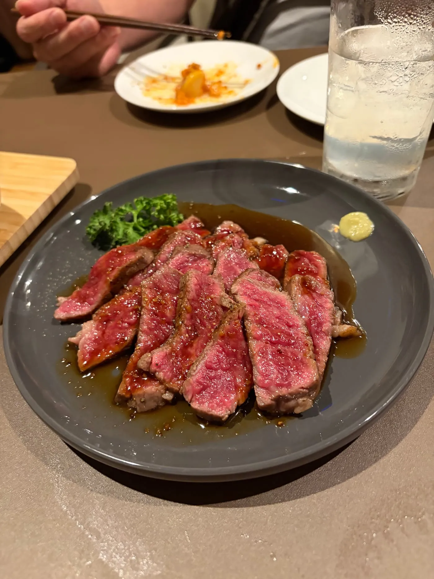 灰色の丸い皿に並んだステーキ。中がピンク色に焼けた肉が五切れほど重なり、まわりに和風のたれがたまっている。奥に薬味、手前にパセリが添えられている
